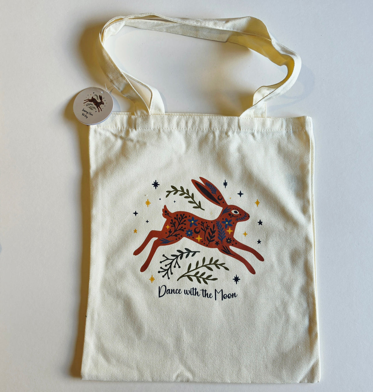 An off-white canvas tote bag with a printed illustration of a hopping hare in red, blue, and green, surrounded by stars and the text 'Dance with the Moon'. A small round tag is attached to one of the handles, and the bag is photographed flat on a light gray background.