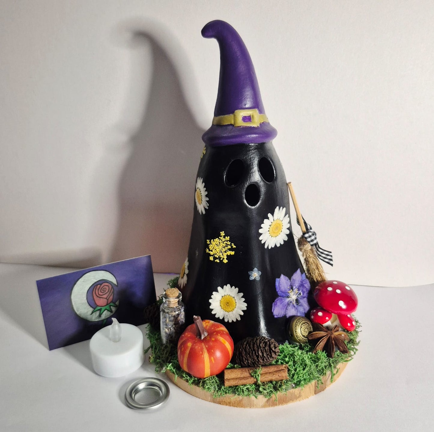 A black decorative Witchy Ghost shaped cone burners on a white surface against a plain wall, with real pressed flowers and magical trinkets with a small LED candle, cone tray and card beside it.