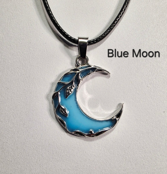 A crescent moon pendant necklace with a light blue enamel center and silver-toned leafy edges, hanging from a black cord on a light gray background. The text 'Blue Moon' appears to the right.