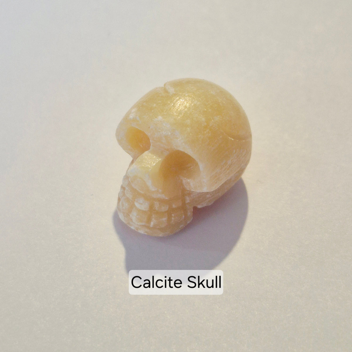 A small carved calcite skull charm in a pale yellowish-beige color, photographed on a light neutral background with a text label reading 'Calcite Skull' underneath.