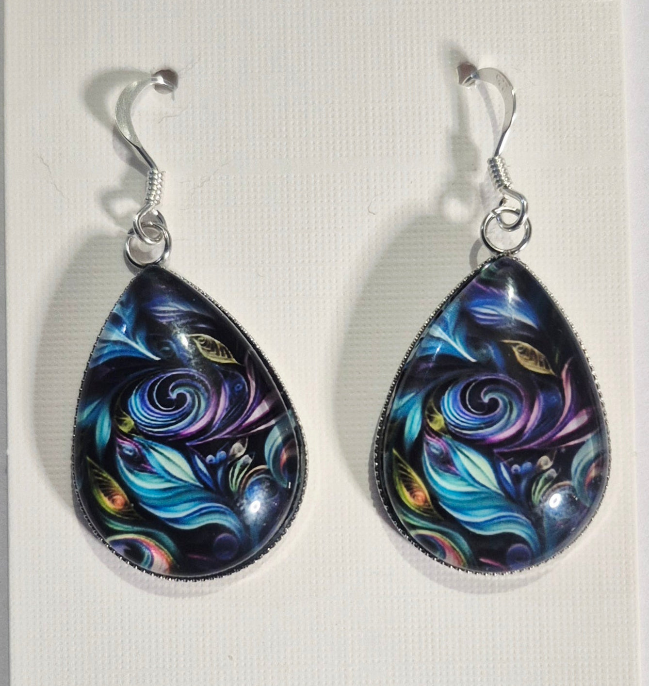 A pair of teardrop-shaped dangle earrings with colorful abstract cosmic swirl designs in blue, purple, pink, and yellow on a dark background, suspended from sterling silver hooks