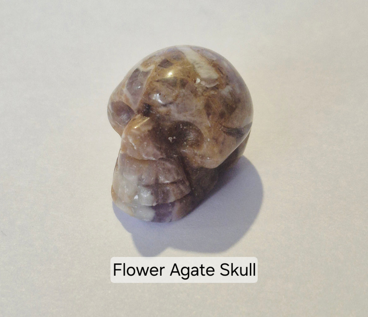 A carved Flower Agate skull figurine with natural banded patterns of brown, beige, and white, displayed on a neutral light gray background with a text label underneath.