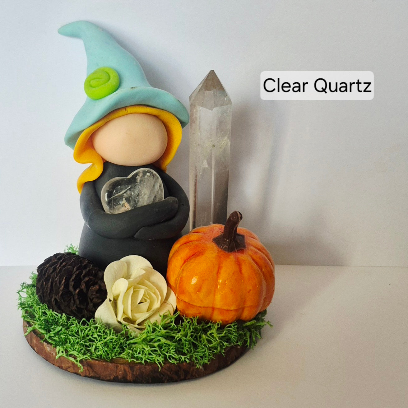A decorative witch figurine with a light blue hat, yellow hair, and a clear quartz crystal held in its hands, displayed on a mossy base with a small orange pumpkin, pinecone, cream rose, and a tall clear quartz crystal tower against a plain white background.