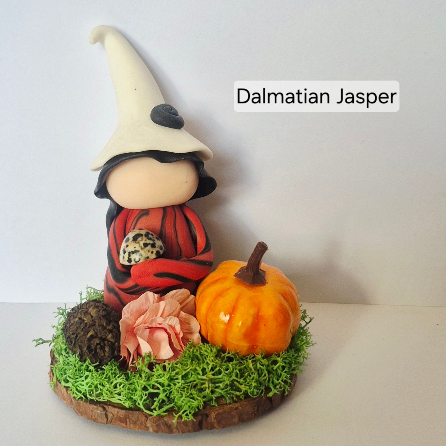 A handmade clay witch figurine labeled 'Dalmatian Jasper' wearing a cream hat and red-and-black striped outfit, posed with a small orange pumpkin, pink flower, pinecone, and green moss on a wooden base against a plain light background.