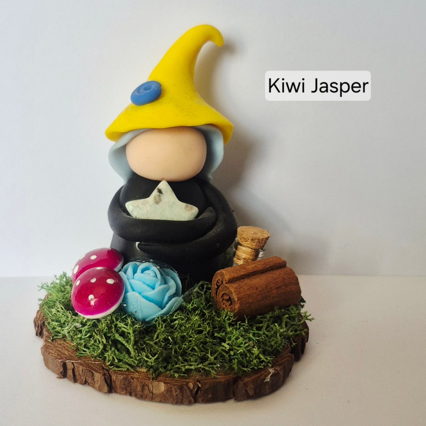 A handmade clay witch figurine labeled 'Kiwi Jasper' sits on a mossy wooden base. The figure wears a bright yellow pointed hat with a blue button and a black robe, holding a gemstone star.