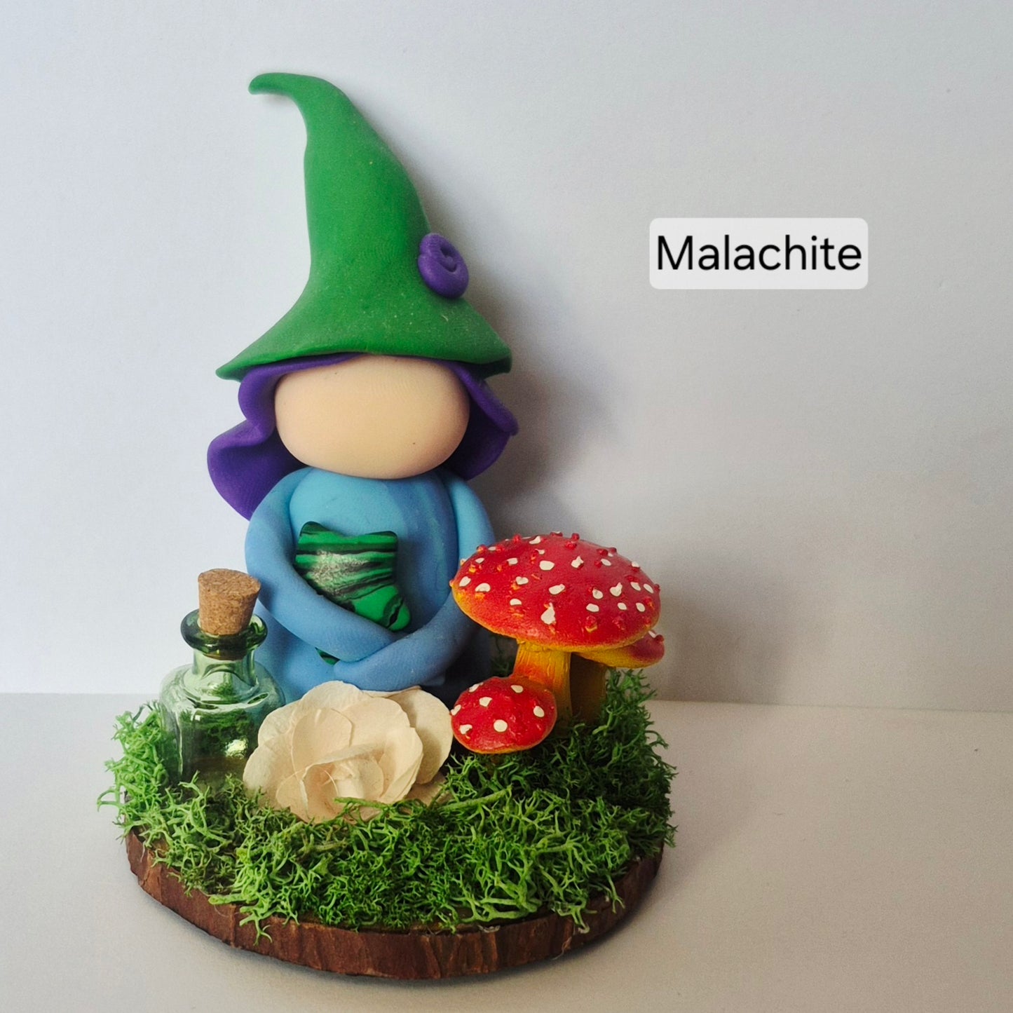 A handmade clay witch figurine with a Malachite star, sitting on a mossy base with red and white mushrooms, a small glass bottle, and a flower. The background is a plain light gray wall.