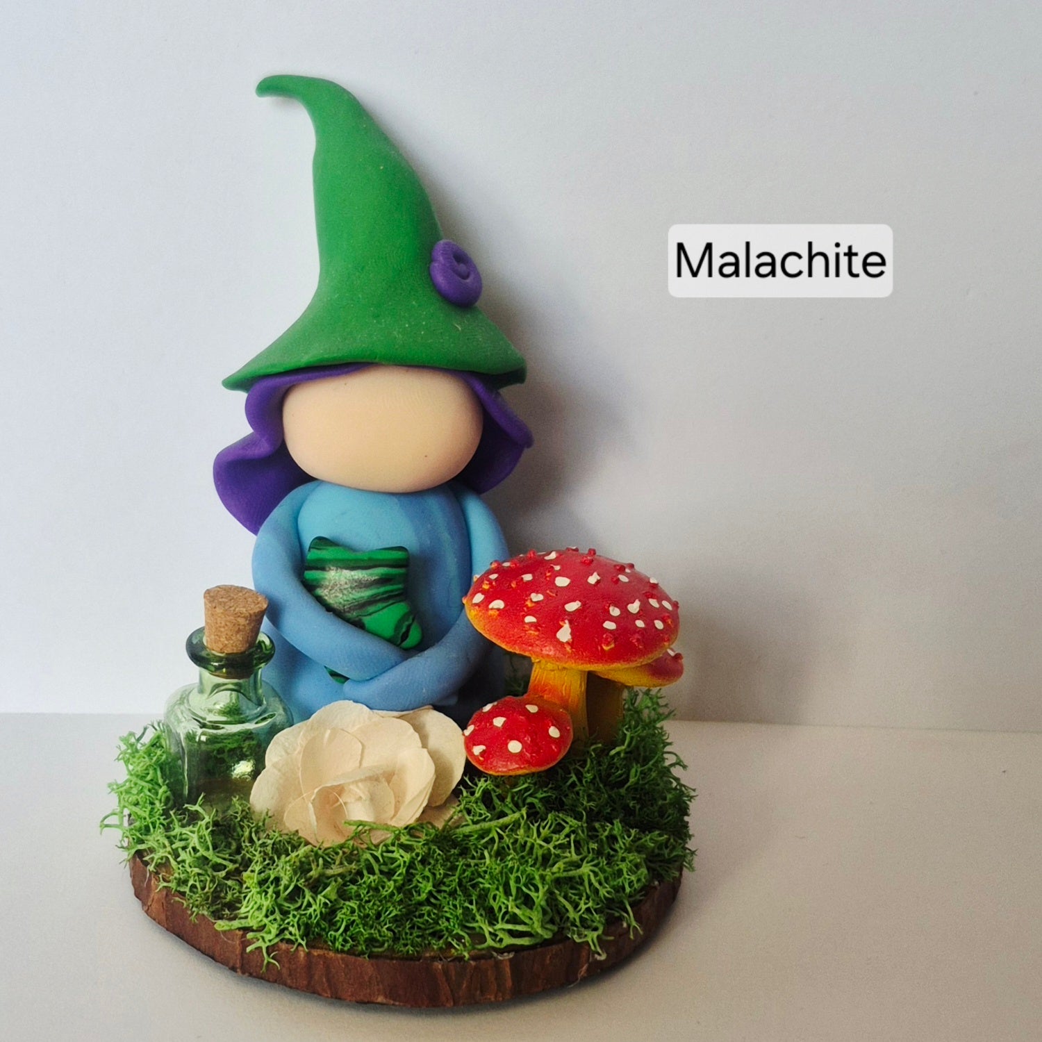 A handmade clay witch figurine with a Malachite star, sitting on a mossy base with red and white mushrooms, a small glass bottle, and a flower. The background is a plain light gray wall.