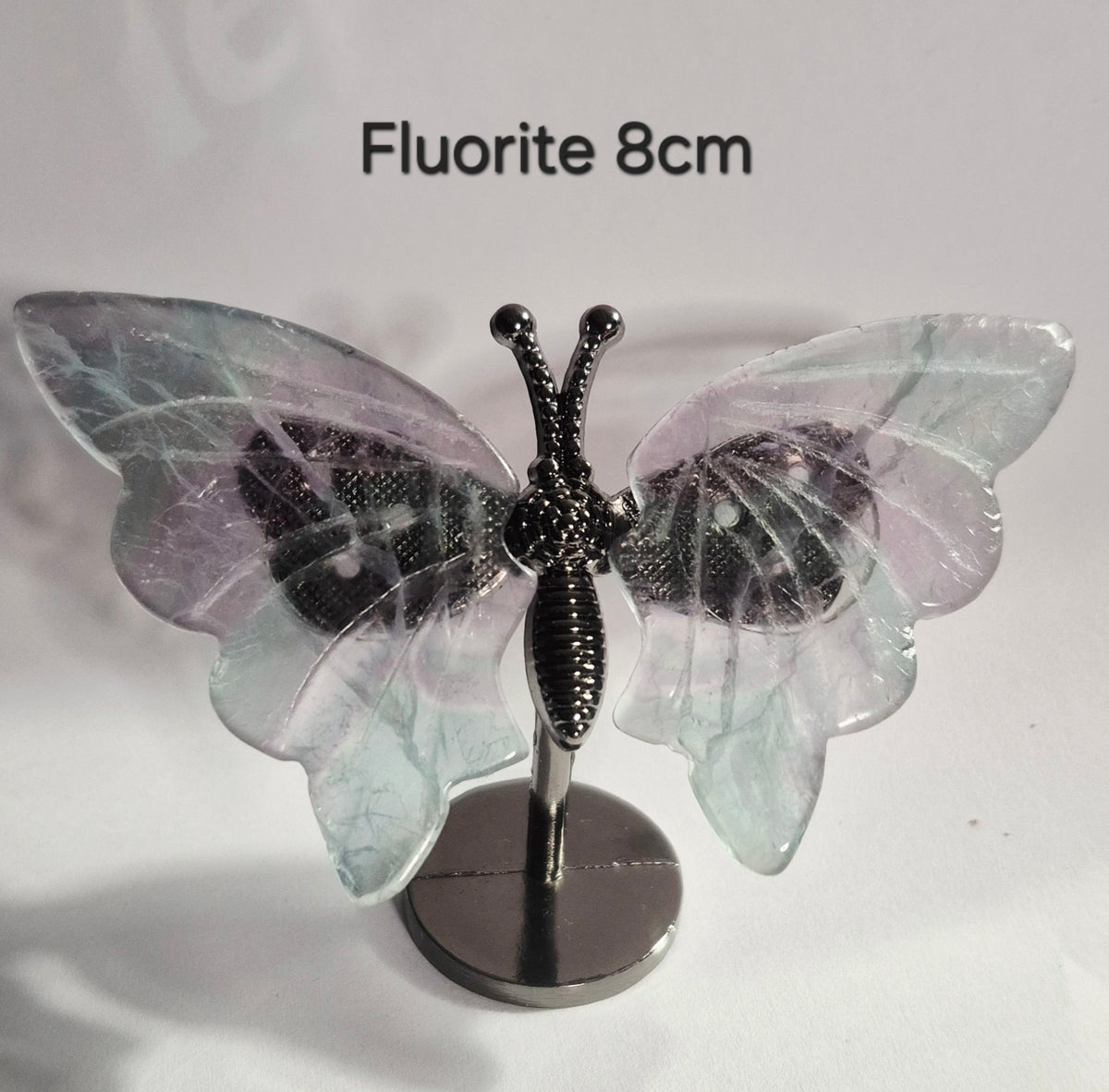 A butterfly-shaped crystal stand made of fluorite with a black metal wire body and stand, displayed on a white background with the text 'Fluorite 8cm' at the top.