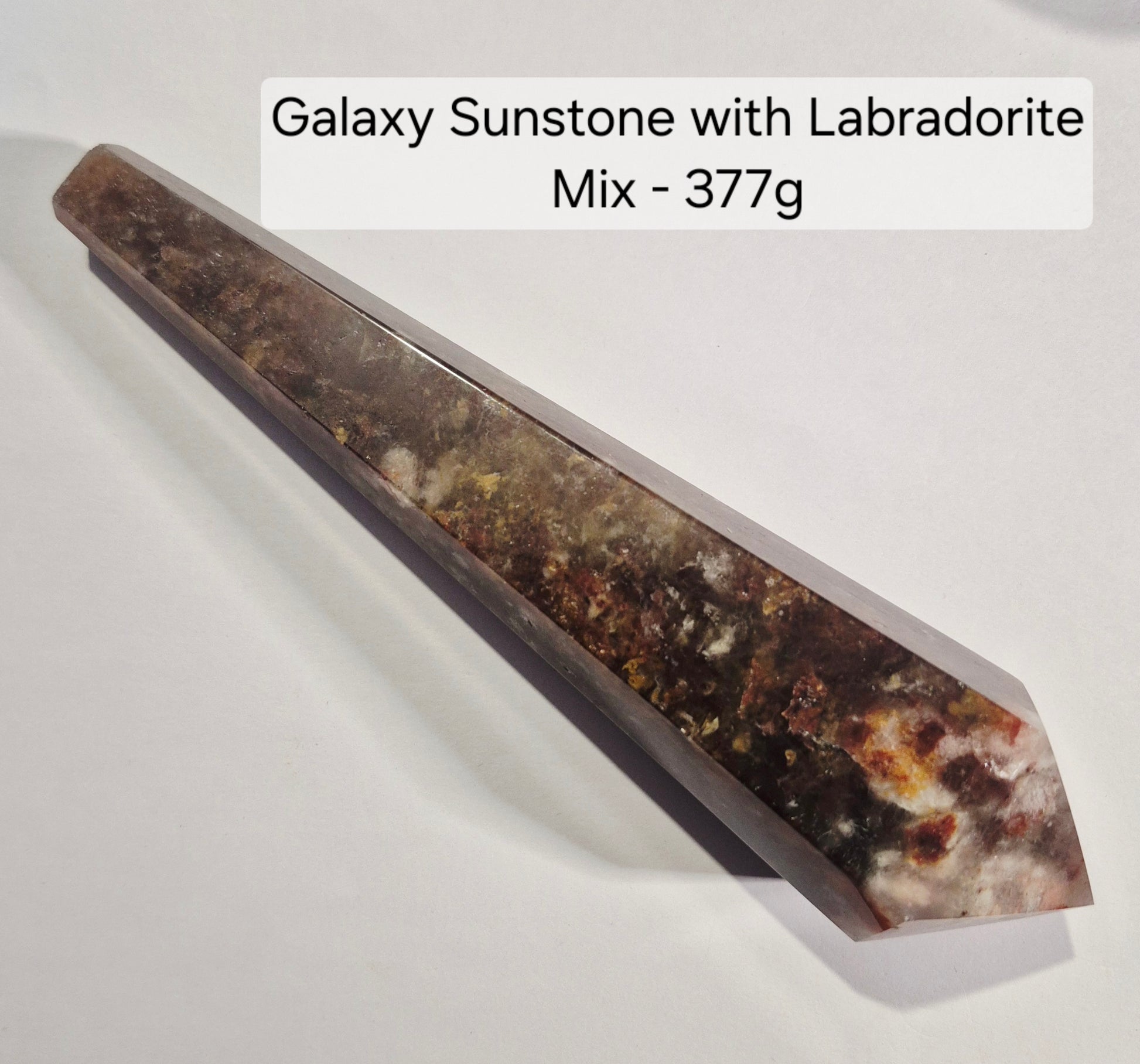 A faceted crystal point labeled 'Galaxy Sunstone with a flashy Labradorite Mix - 377g' on a plain light background. The stone shows a mottled mix of brown, gray, black, and white crystalline patterns.