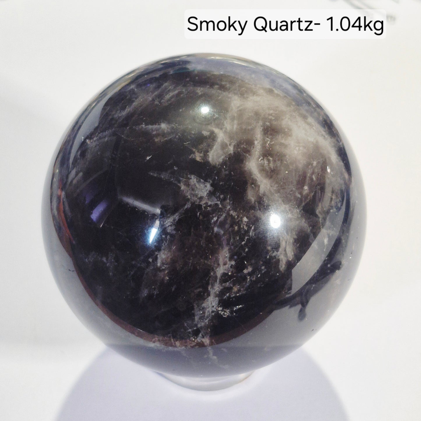 A polished spherical Smoky Quartz crystal ball with dark gray to black smoky patterns and lighter gray veining, displayed on a white surface with a text overlay reading 'Smoky Quartz- 1.04kg' at the top right.