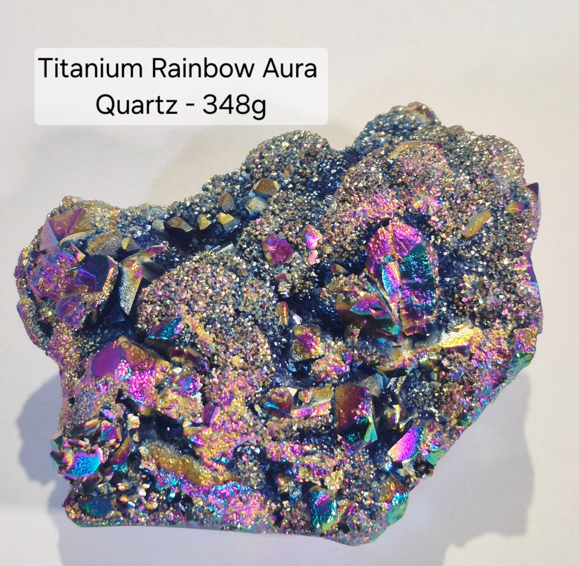 A raw mineral specimen labeled 'Titanium Rainbow Aura Quartz - 348g' showing highly iridescent, metallic crystal structures with a mix of gold, purple, blue, and pink hues on a light gray background.
