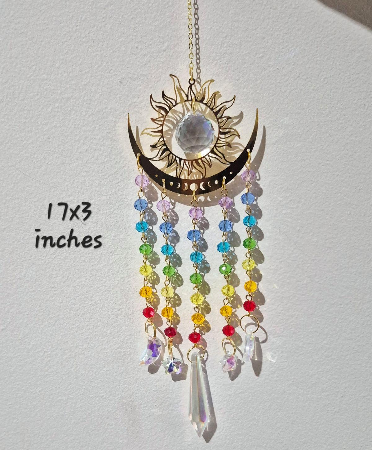 A gold-toned crescent moon and sun hanging ornament with a large clear stone in the sun face and dangling rainbow beaded strands, shown against a white wall with handwritten text reading 17x3 inches.