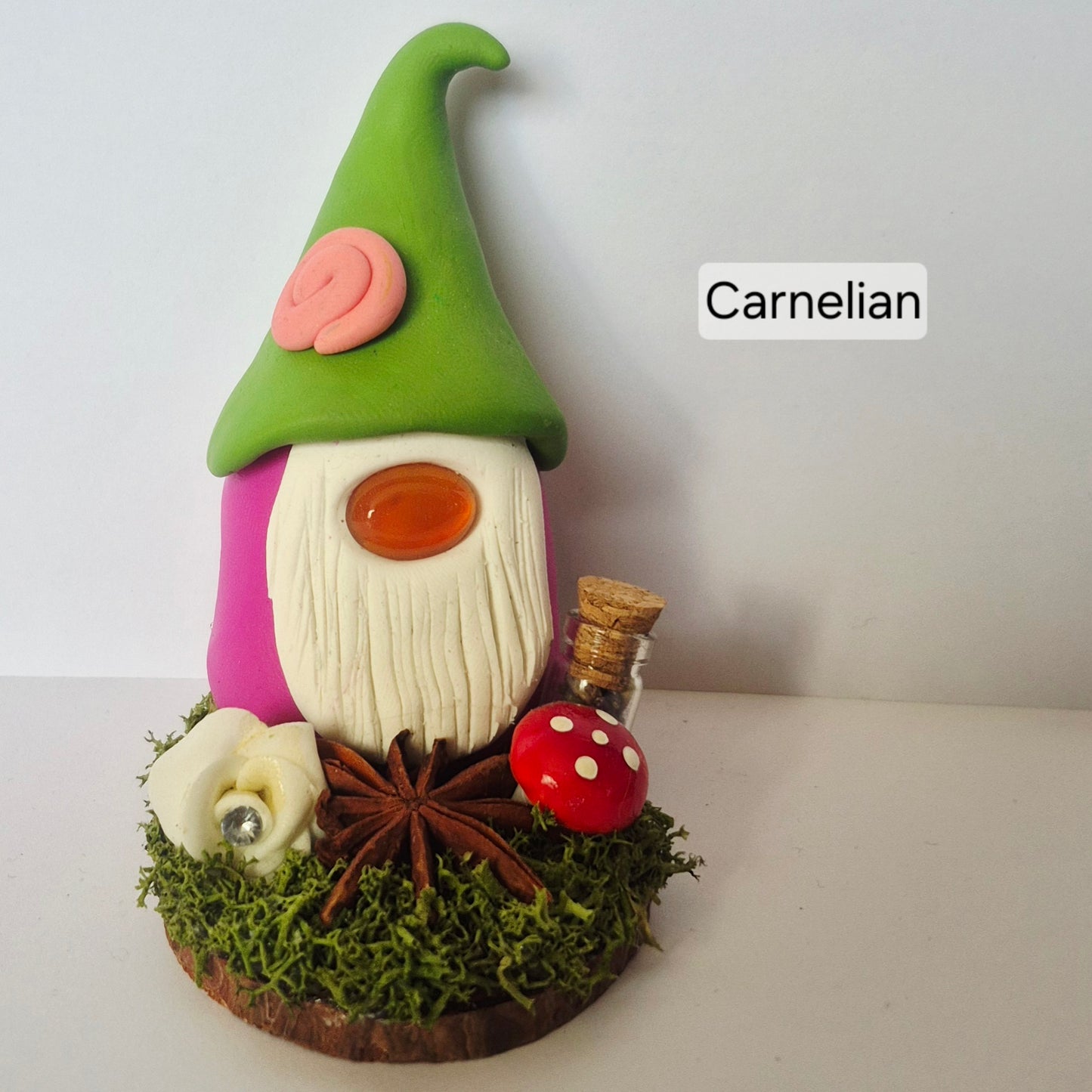 A handmade clay gonk figurine with a green pointed hat, pink body, and white beard, displayed on a mossy base with a cinnamon stick, star anise, a red mushroom, and a small bottle. The text 'Carnelian' is displayed on the right.