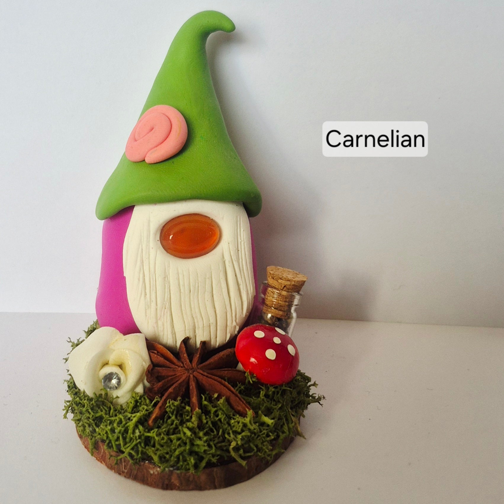 A handmade clay gonk figurine with a green pointed hat, pink body, and white beard, displayed on a mossy base with a cinnamon stick, star anise, a red mushroom, and a small bottle. The text 'Carnelian' is displayed on the right.