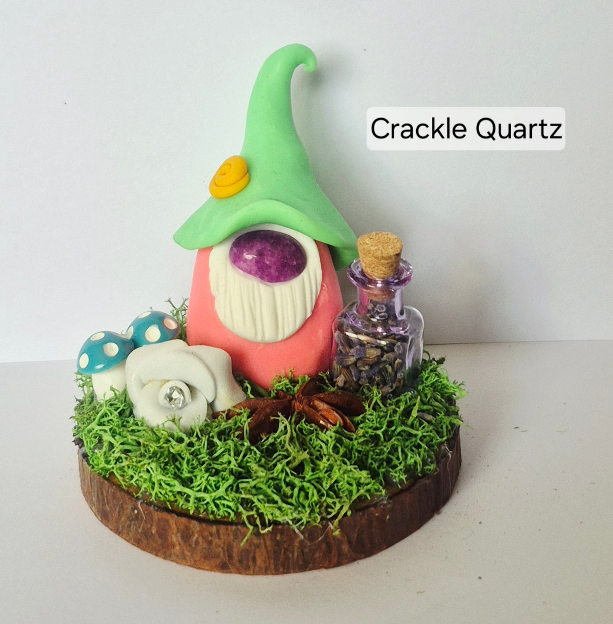 A handmade clay gonk figurine sitting on a mossy base with a small glass bottle, mushrooms, star anise and a flower set against a plain white background. The text 'Crackle Quartz' is visible in the upper right.