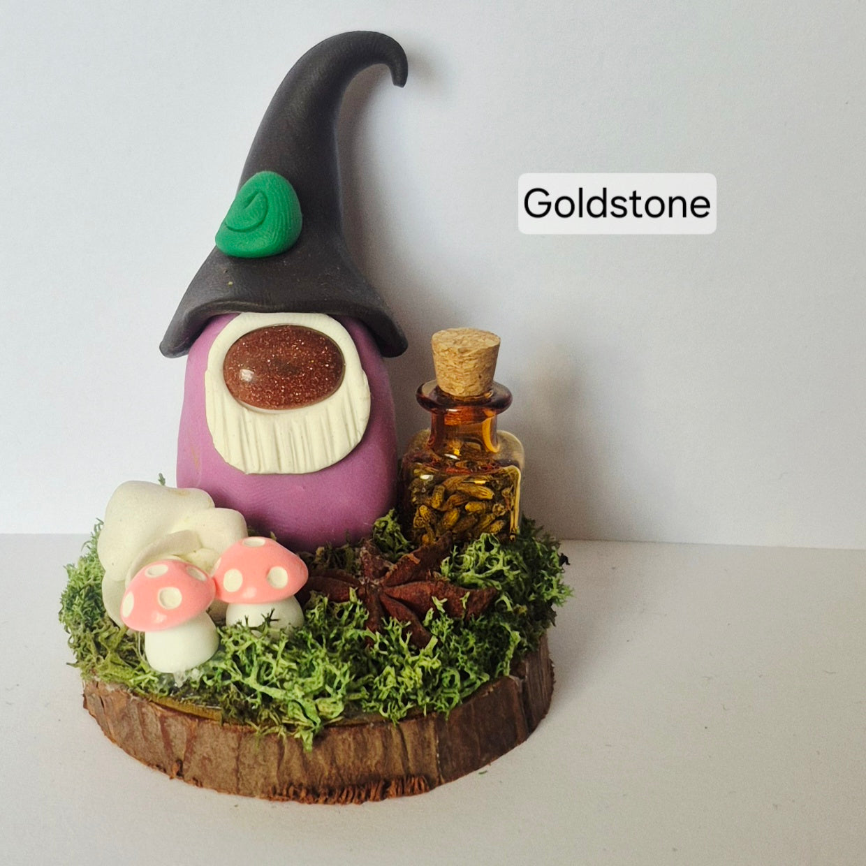 A small handmade decorative gonk figurine with a black pointed hat, purple body, white beard, pink and white mushrooms, green moss, and a small amber bottle on a wood slice base. The word 'Goldstone' appears in a white label near the top right.