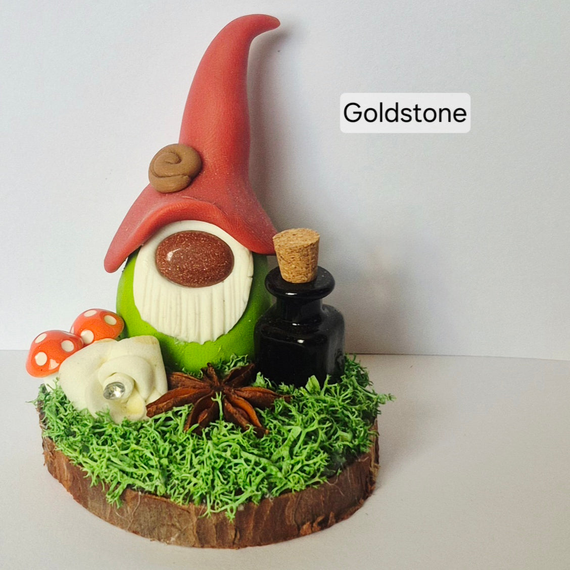 A decorative gonk figurine on a wood slice base with green moss, a small black bottle, mushrooms, star anise, and a white rose. The hat is a matte red and the text 'Goldstone' appears near the top right.
