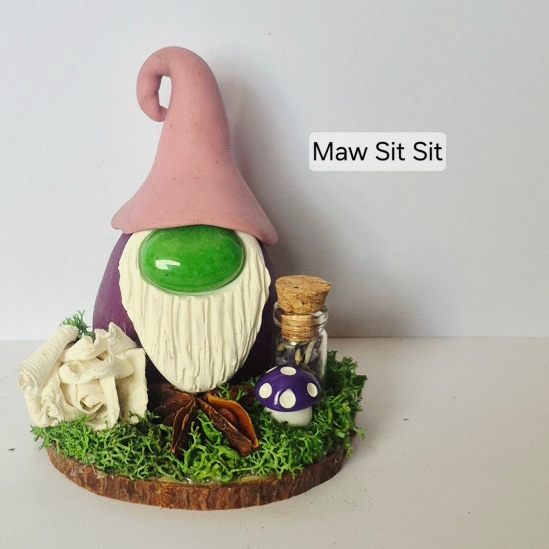 A handmade clay gonk figurine with a pink hat, white beard, and purple body, displayed on a mossy base with a small jar and mushroom. A text overlay reads 'Maw Sit Sit'.