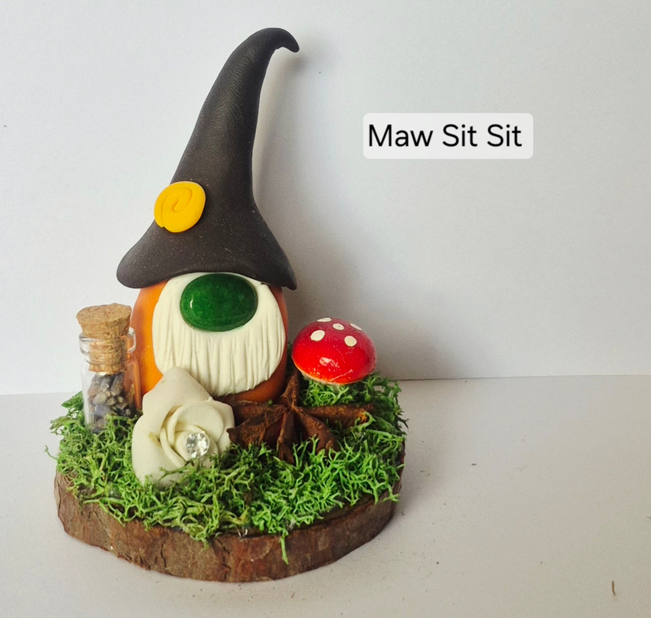 A handmade clay gonk figurine sitting on a small mossy base, surrounded with a jar, mushroom, white flower and star anise. The gonk has a tall black hat with a yellow button, a white beard, and gemstone nose.