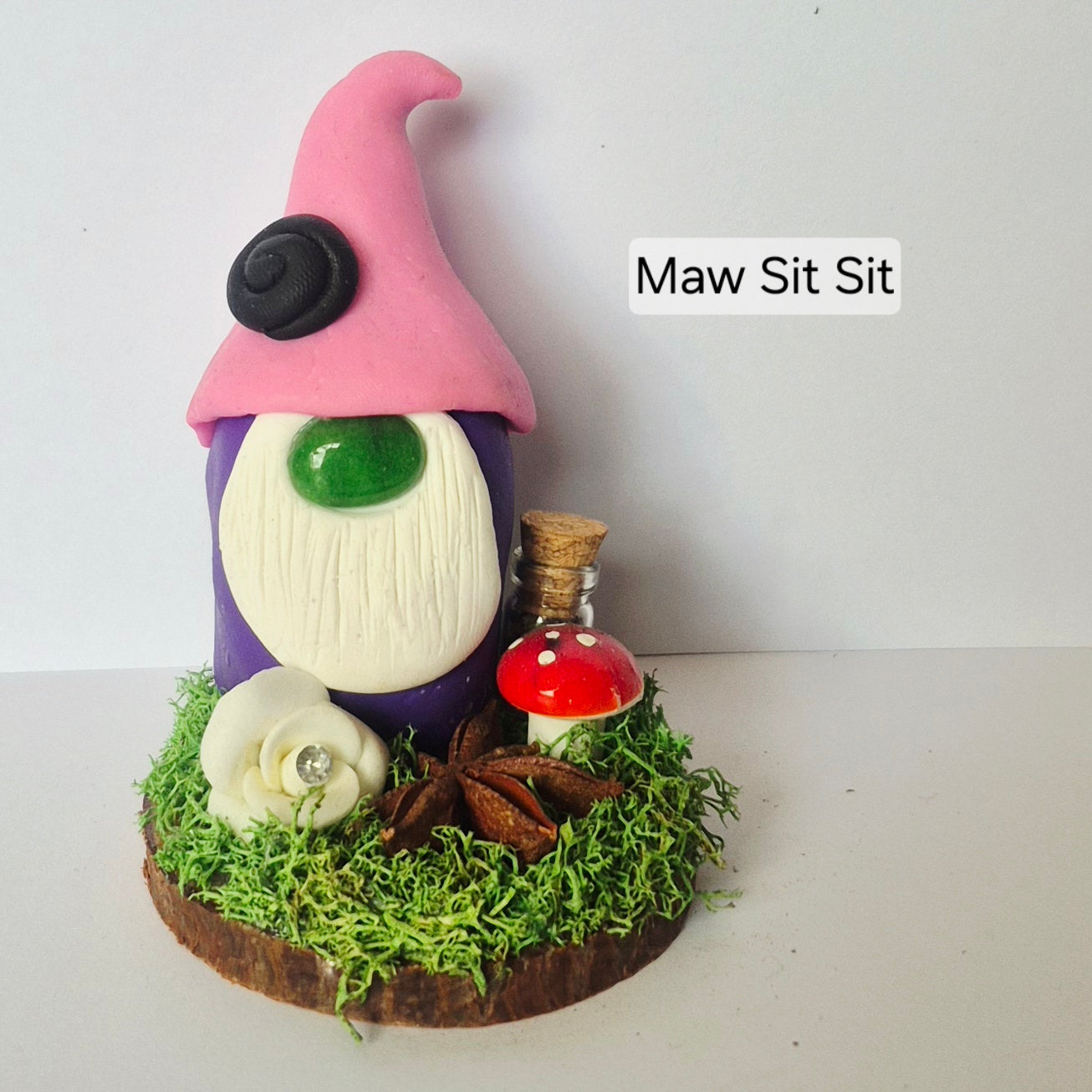 A handmade ceramic gonk figurine with a pink hat, purple body, white beard, gemstone nose, and a small red-and-white mushroom on a mossy base, with a jar, flower and star anise. Photographed against a plain light background. The text 'Maw Sit Sit' appears to the right.