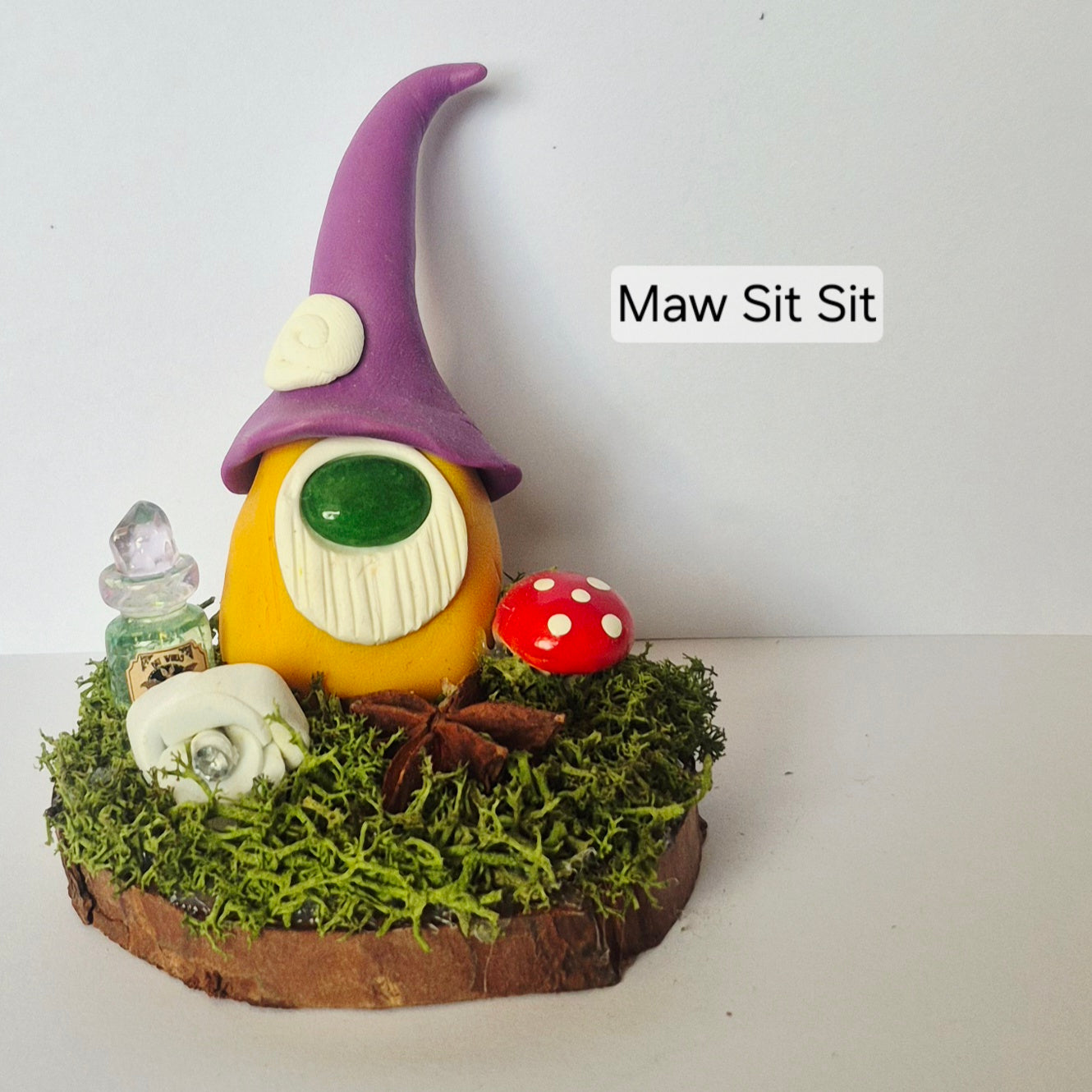A small decorative gonk figurine sitting on a mossy log base. The gonk has a purple pointed hat with a white heart detail, a yellow-orange body, a white beard, a gemstone nose, and is accompanied by a red-and-white mushroom, a small bottle, and a white flower.