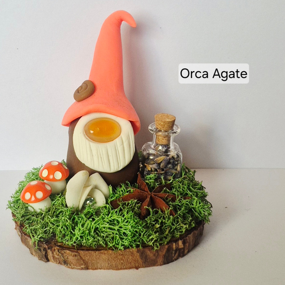 A handmade miniature gonk figurine displayed on a decorative mossy base with small mushrooms, a potion jar, flower and star anise on a wood slice. The gonk has a bright coral-pink hat, a white beard, and a brown body.