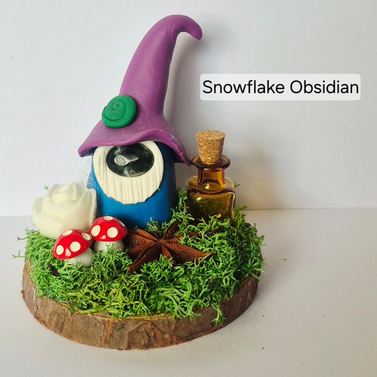 A handmade gonkfigurine crafted from blue resin, a purple hat, and a white beard, displayed on a mossy wooden base with red and white mushrooms, a white rose, star anise and a small amber bottle. The text 'Snowflake Obsidian' is visible on the right.