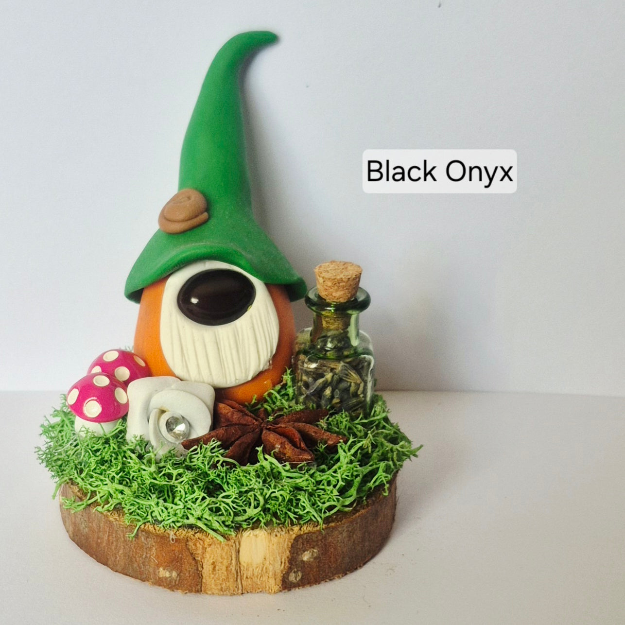 A handmade miniature gonk figurine made of polymer clay, sitting on a small wooden slice base with artificial moss, a small glass bottle, pink mushrooms, and star anise. The text 'Black Onyx' is displayed on the right side.