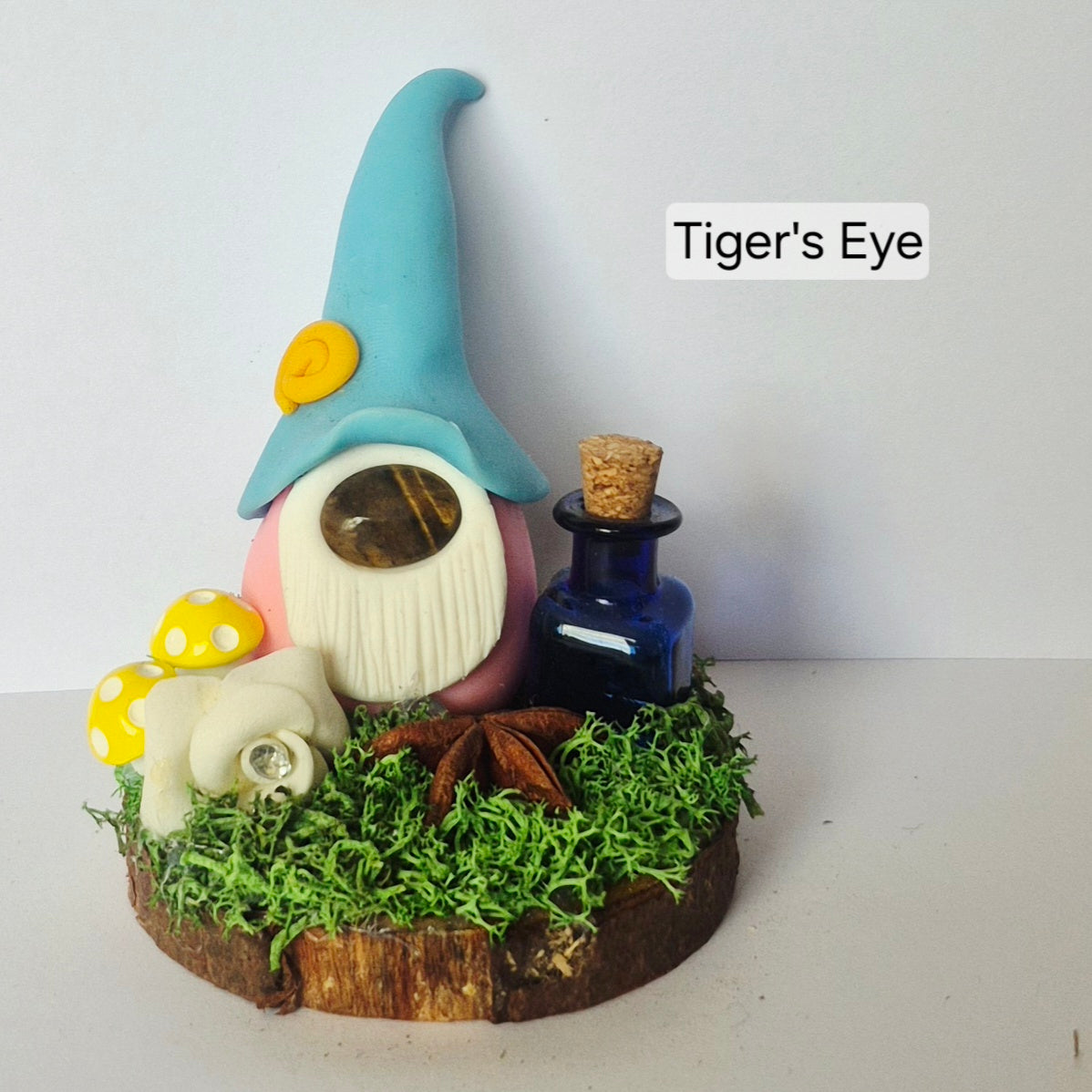A handmade clay gonk figurine with a tall blue hat, white beard, pink body, and yellow accent, displayed on a wood slice base with faux moss, a small blue bottle, mushrooms, and a white flower. The text 'Tiger's Eye' is visible in the upper right.