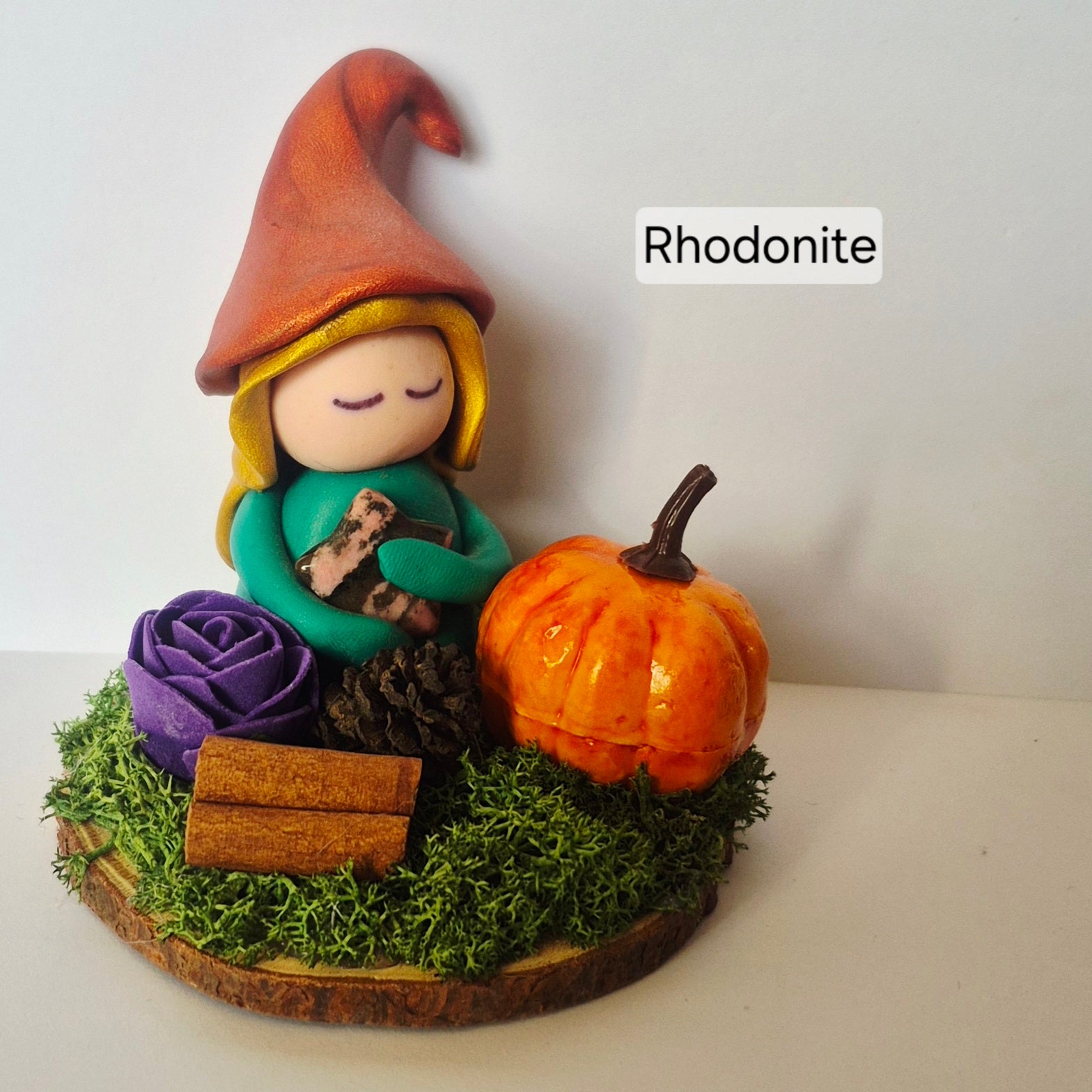 A handmade clay figurine of a witch sitting next to a small orange pumpkin, displayed on a mossy wooden base with a purple rose and cinnamon sticks. The figurine is labeled 'Rhodonite' in a text overlay.