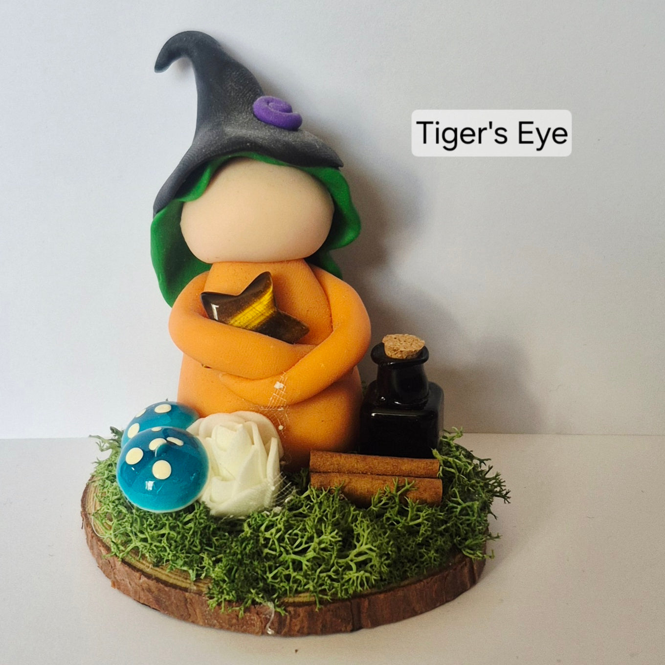 A handmade clay figurine of a witch with a pointed black hat and green hood, holding a yellow book, standing on a mossy wooden base with a blue mushroom, white rose, cinnamon sticks, and a small black bottle. The text 'Tiger's Eye' is displayed on the right.