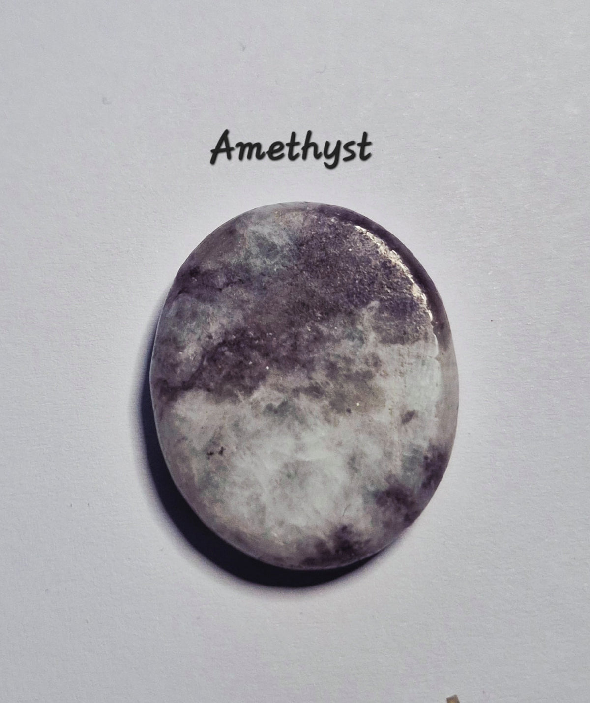A polished oval amethyst gemstone with natural purple and white marbled patterns, displayed on a light gray background with the word 'Amethyst' printed above it.