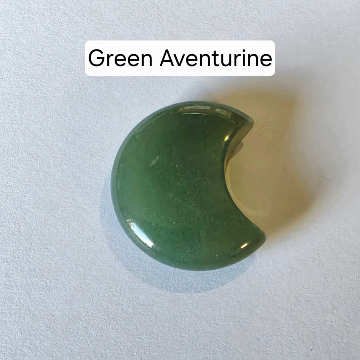 A polished crescent-shaped green aventurine stone on a textured white background with a text label reading 'Green Aventurine' at the top.