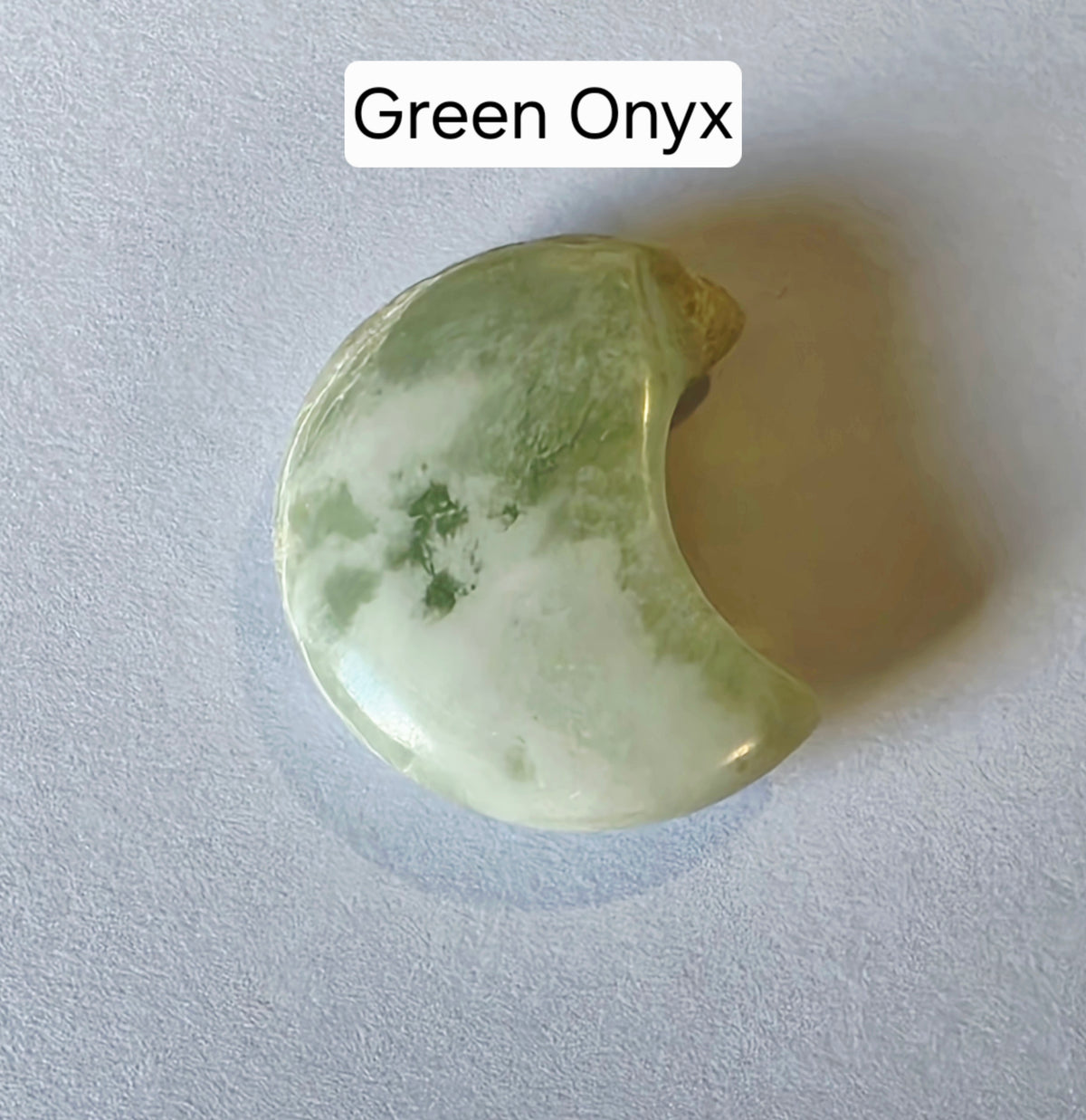 A crescent-shaped green onyx stone with natural marbled green and pale cream patterns, displayed on a textured light gray background with a text label reading 'Green Onyx' at the top.