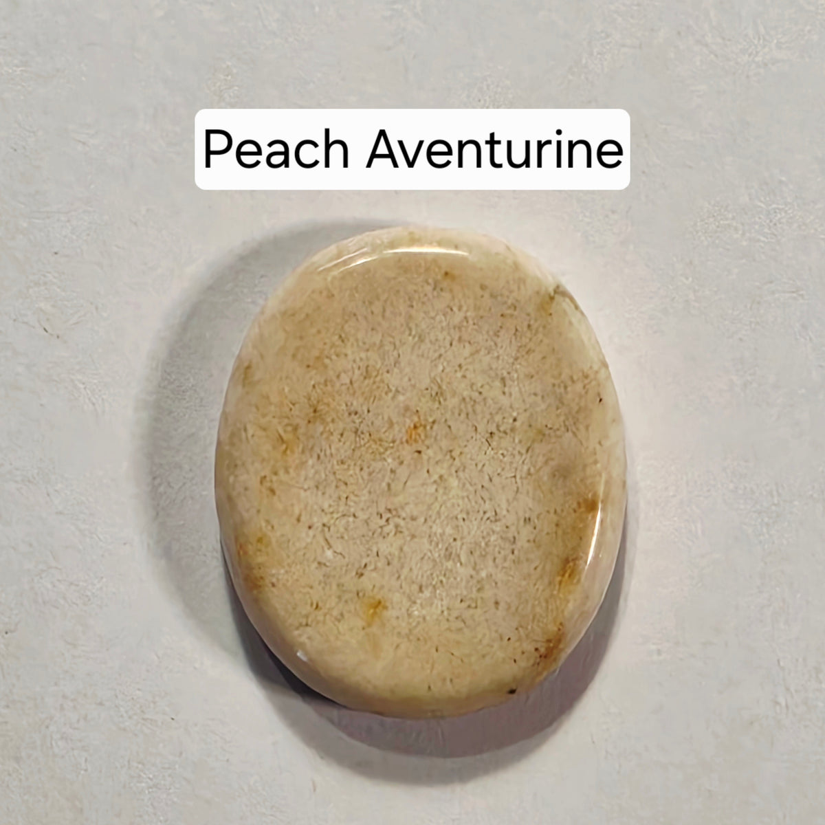 A polished peach aventurine worry stone with a smooth, rounded oval shape, displayed on a textured light gray background with a text label reading 'Peach Aventurine'.