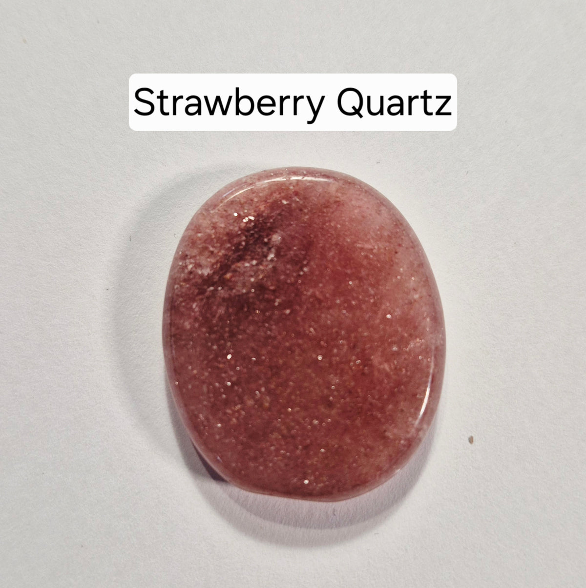 A polished oval worry stone of strawberry quartz gemstone on a light gray background with a text label reading 'Strawberry Quartz' at the top.