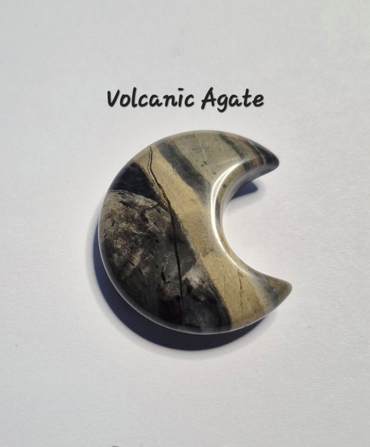 A crescent moon-shaped polished gemstone with a volcanic agate pattern of black, gray, and beige, displayed on a white background with the text 'Volcanic Agate' above it.