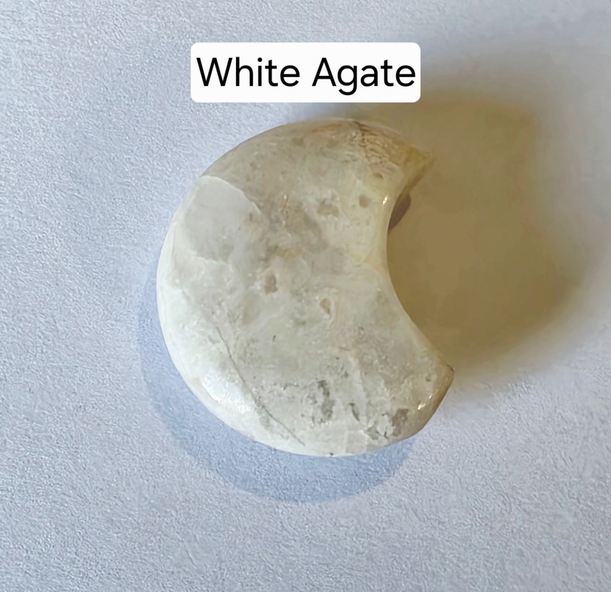A polished crescent moon-shaped white agate stone with natural marbled textures, displayed on a light gray textured background with a text label reading 'White Agate' at the top.