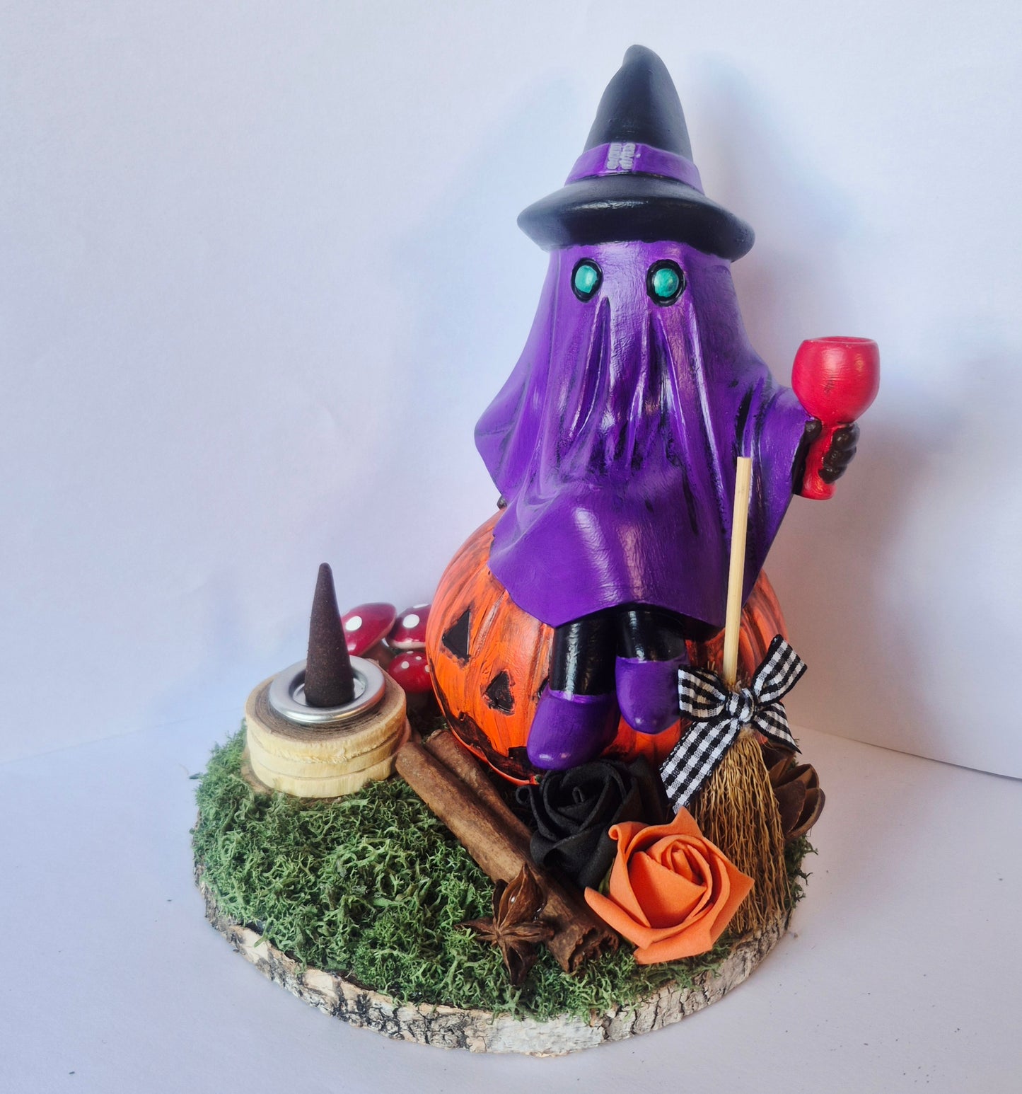 A Halloween-themed decorative figurine featuring a ghost-like witch in a purple cloak and black hat, seated on an orange pumpkin. The figure holds a red goblet and stands on a mossy circular base with a broom, cinnamon sticks, a black rose, an orange paper rose, and small red mushrooms.