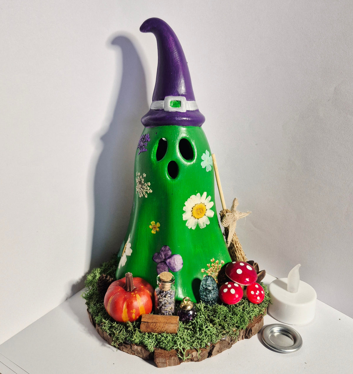 A handmade decorative ghost with a purple witch hat, green body, and Halloween-themed accessories including a small pumpkin, mushrooms, jars, and mossy base, shown on a white surface against a white wall. For LED Candles and Cone burning