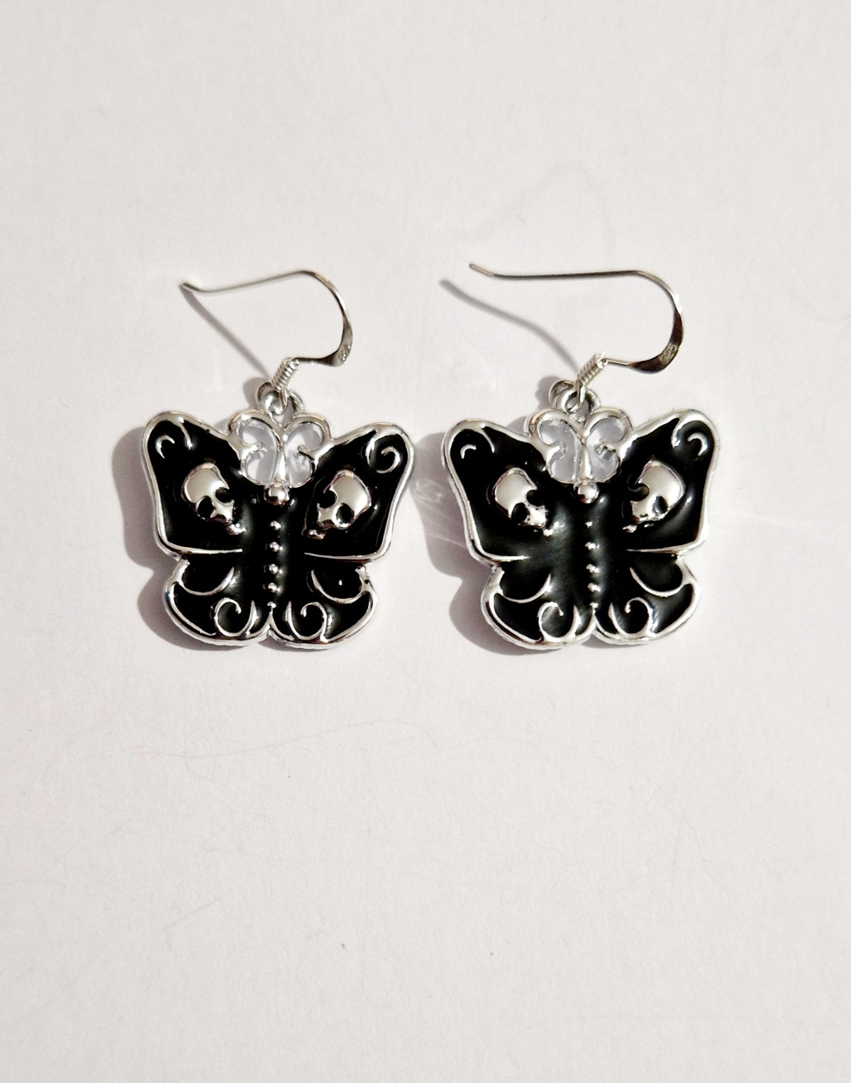 A pair of gothic-style butterfly drop earrings featuring black enamel wings with silver skull details, sterling silver hooks, and clear rhinestone accents, displayed on a light off-white background.