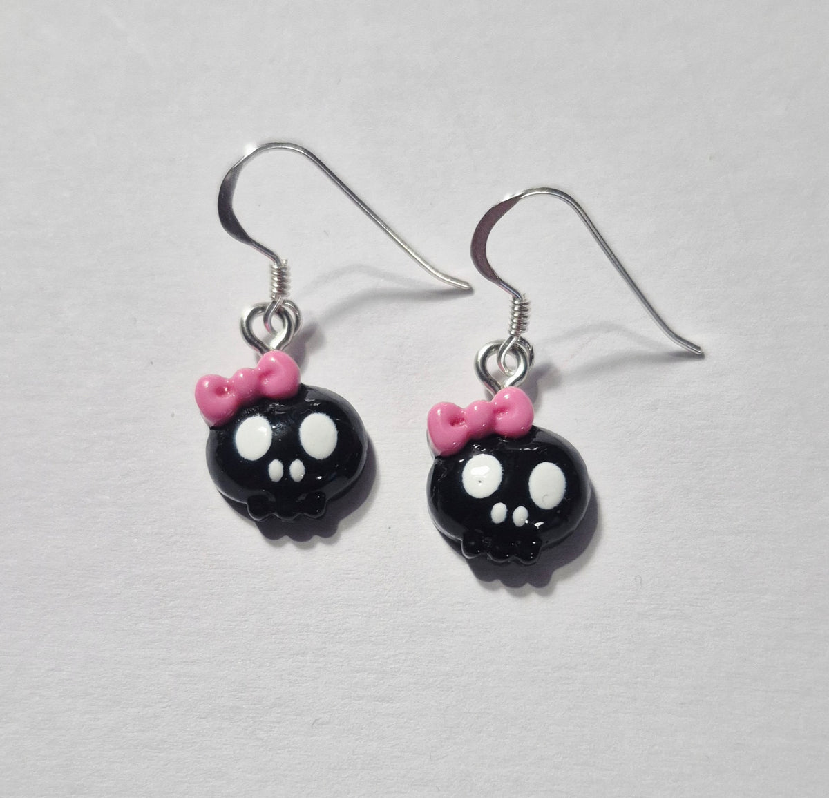 A pair of dangling skull earrings with black glossy skulls, white facial details, pink bows, and sterling silver fishhook wires, shown on a plain light background.
