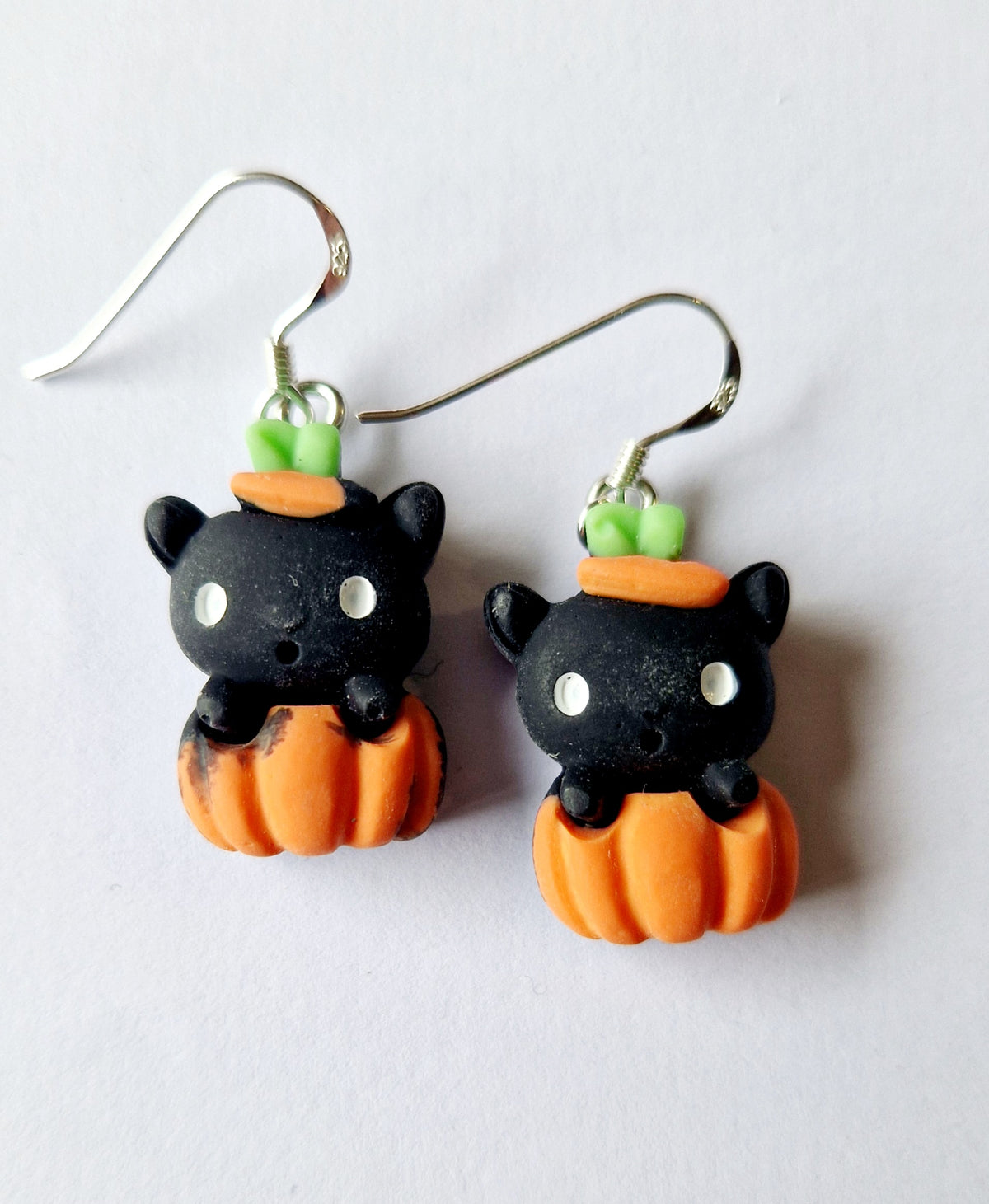 A pair of dangling earrings featuring cute black cat charms sitting in orange pumpkins, with small green leaf accents and sterling silver fishhook wires, shown on a plain light background.