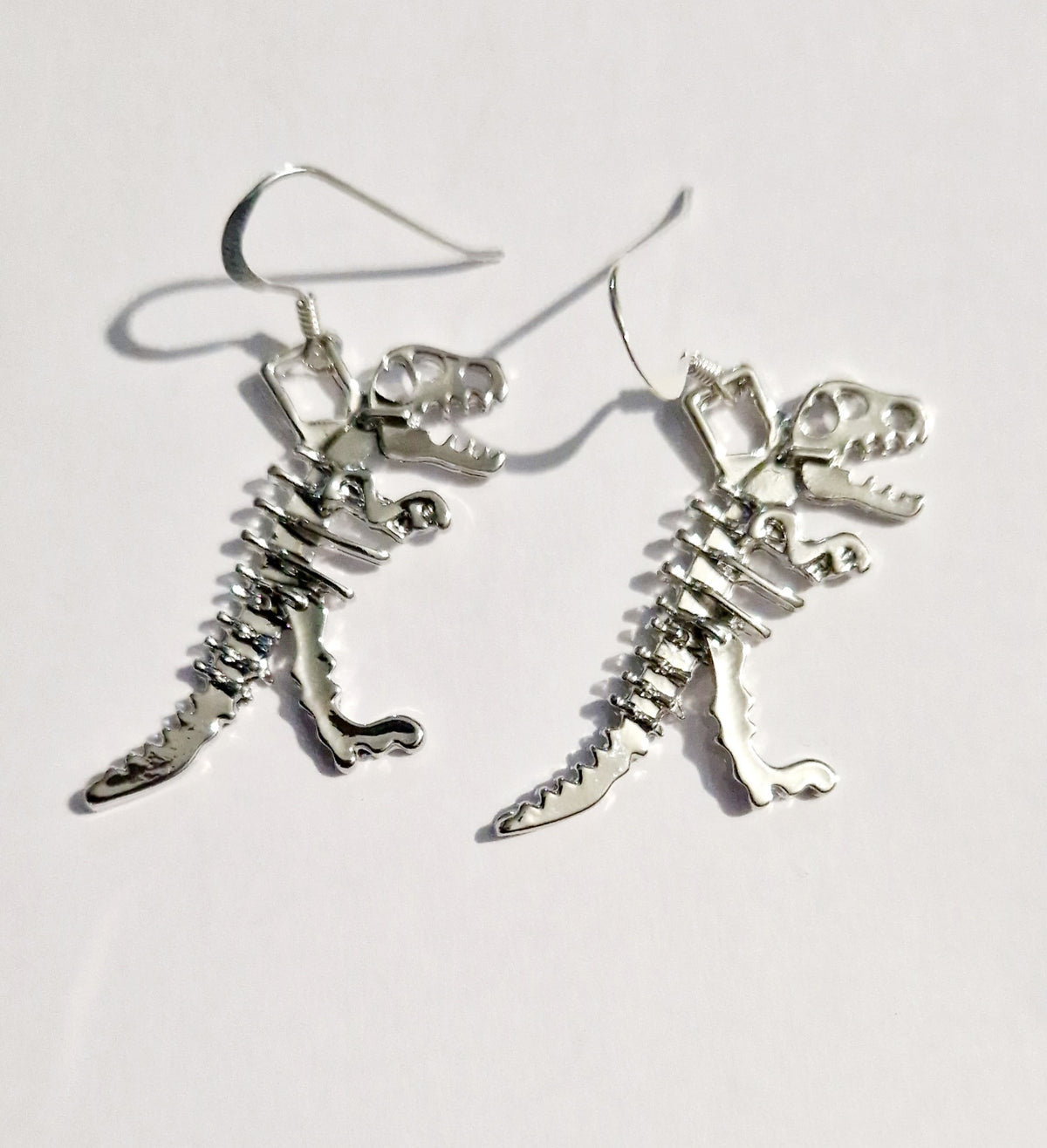 A pair of silver dangling earrings shaped like dinosaur skeletons, photographed on a plain white background.