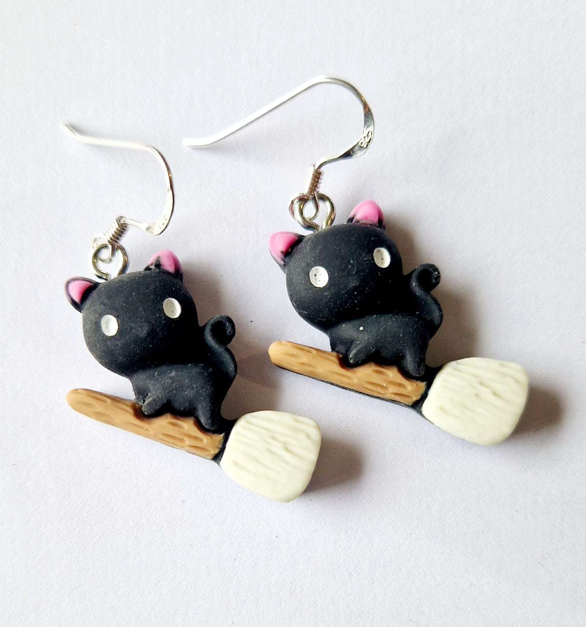 A pair of dangling earrings featuring small black cat charms with pink ears, white eyes, and sterling silver fishhook wires, each attached to a wooden-handled broom with off-white bristles, shown on a plain light background.