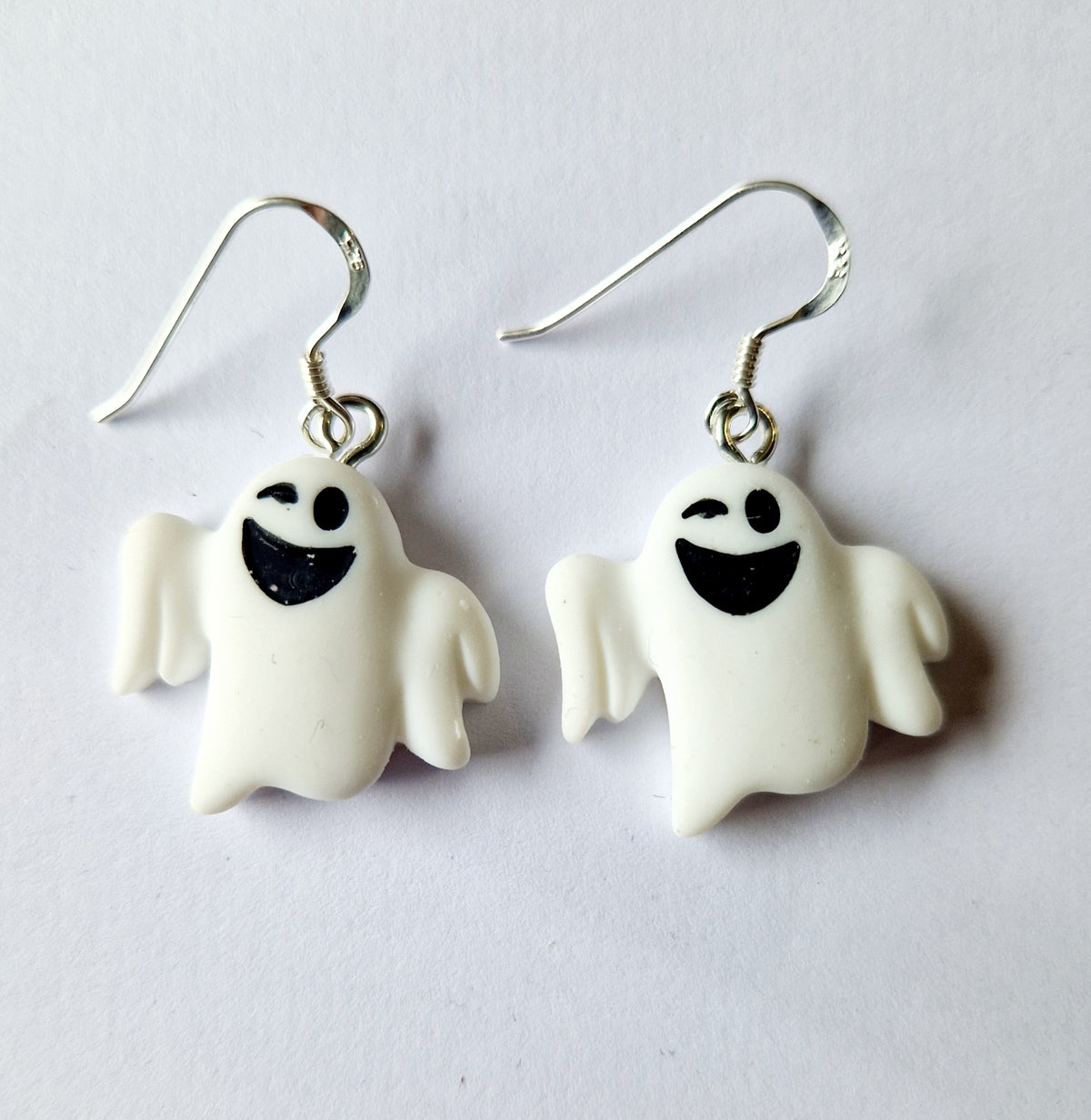 A pair of white ghost-shaped dangle earrings with black smiley faces and sterling silver fishhook wires, shown on a plain light background.
