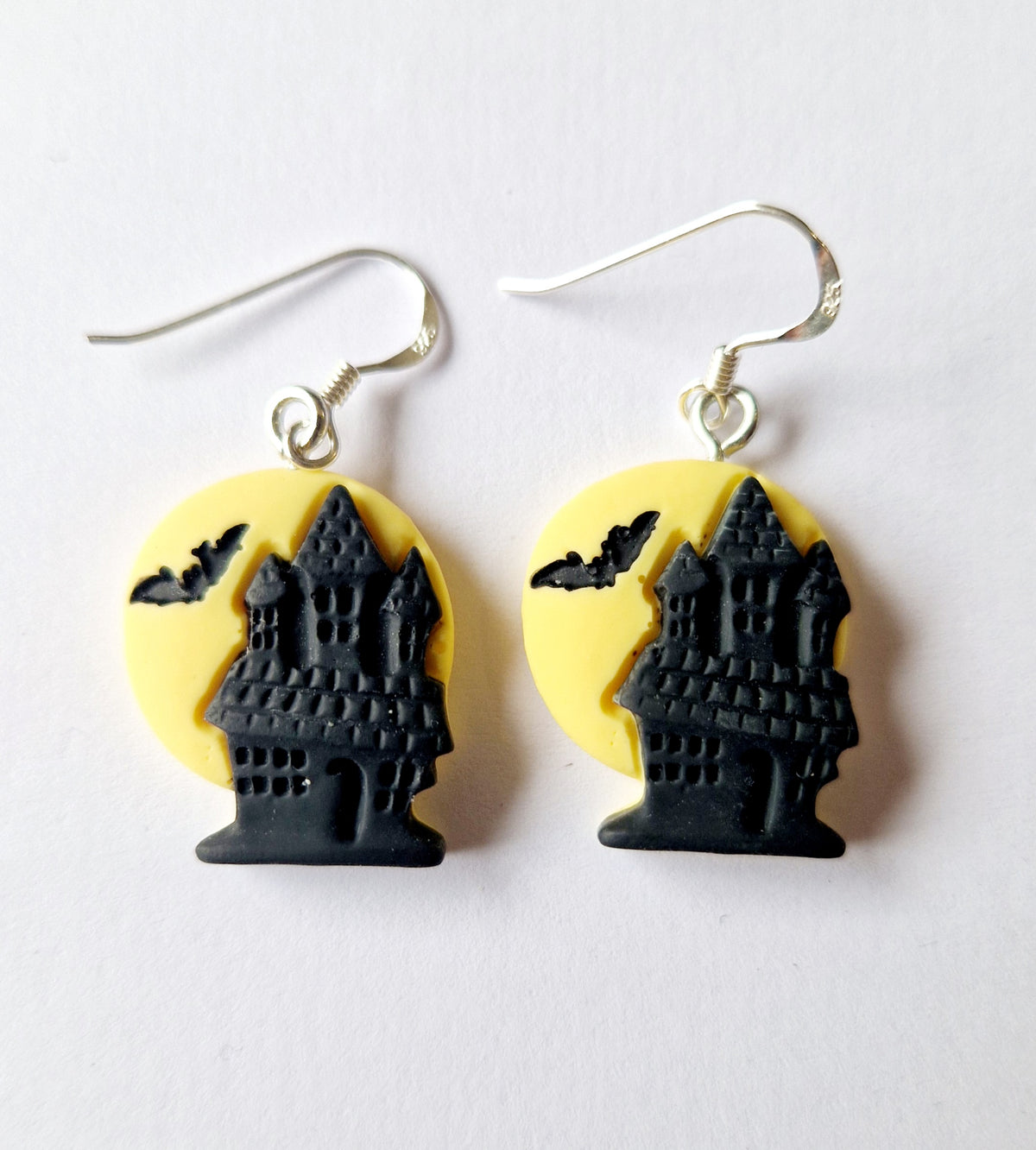 A pair of Halloween-themed dangle earrings featuring a black castle silhouette in front of a yellow circular background, suspended from sterling silver fishhook wires, shown on a plain white surface.
