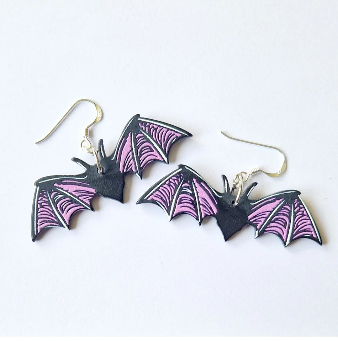 A pair of bat-shaped dangle earrings with black bodies, pink and purple patterned wings, and sterling silver fishhook wires, displayed on a clean white background.