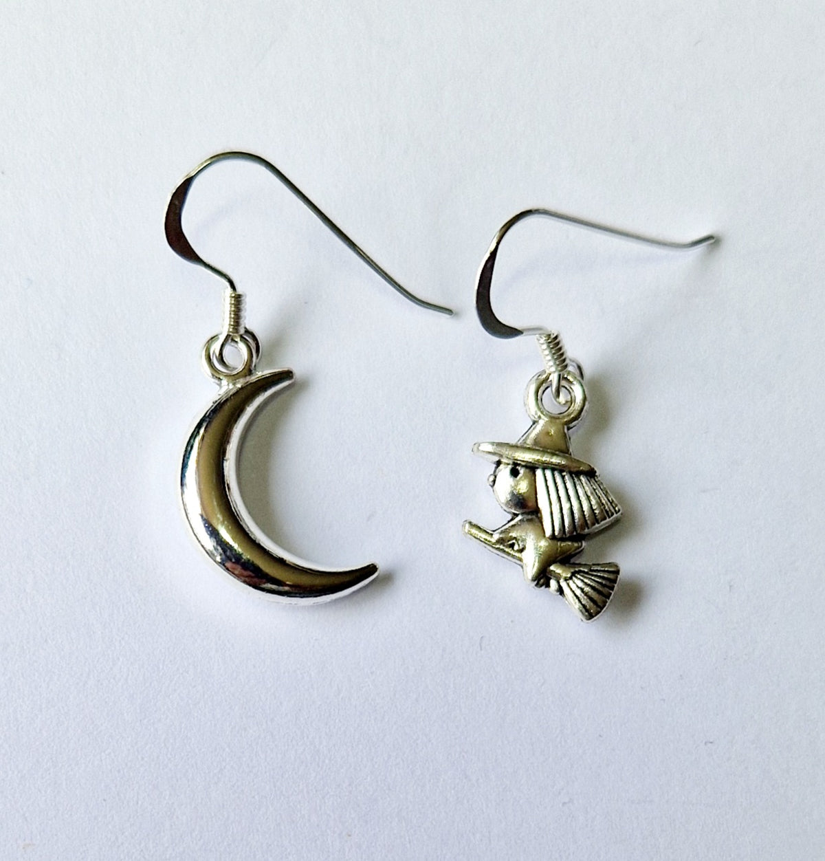 A pair of dangling earrings on a plain white background. One earring features a polished silver crescent moon charm, and the other features an antiqued silver witch riding a broomstick charm.