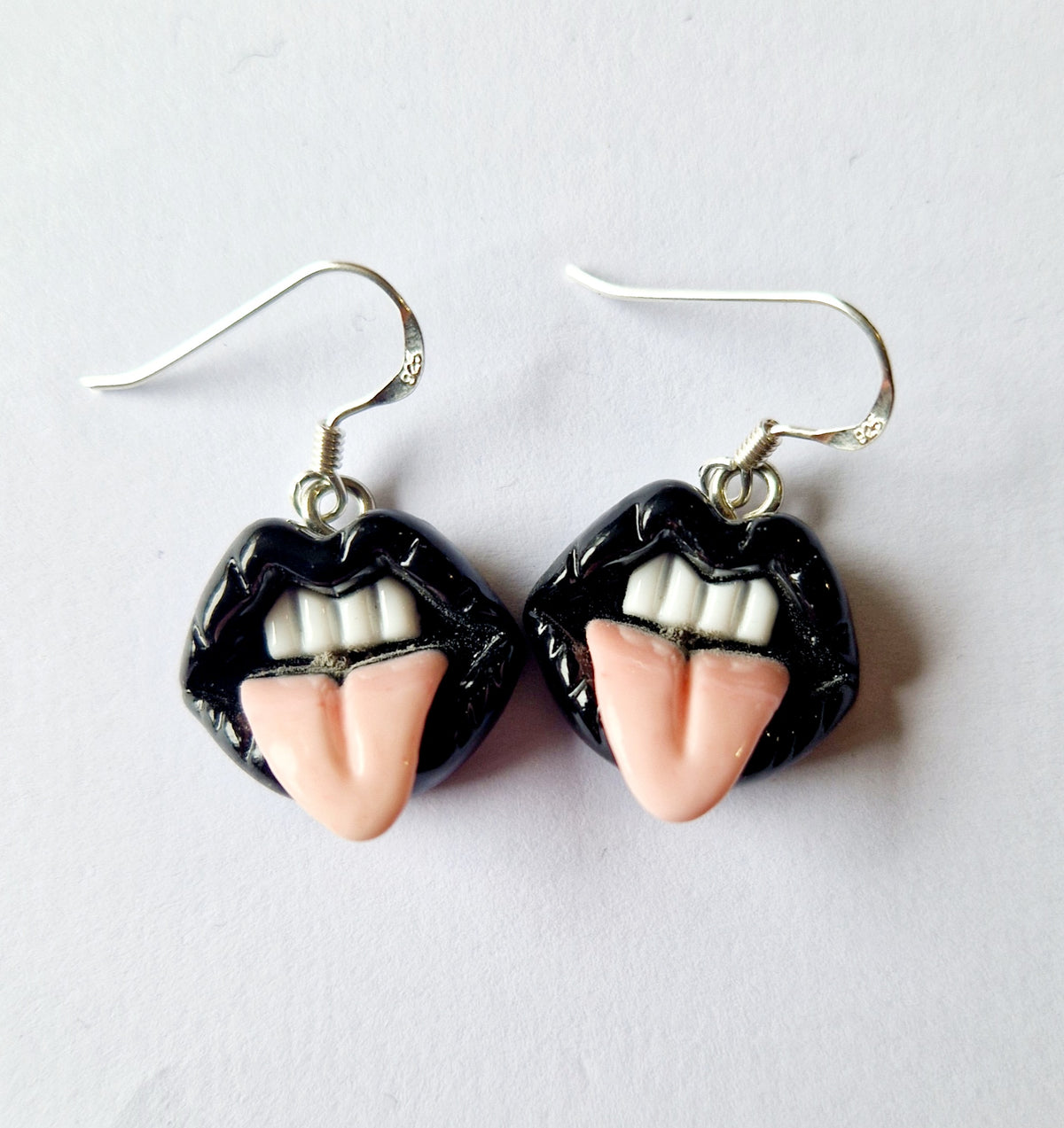 A pair of novelty drop earrings shaped like lips with black glossy lips, white teeth, and pink tongues, hanging from sterling silver fishhook wires on a plain light background.
